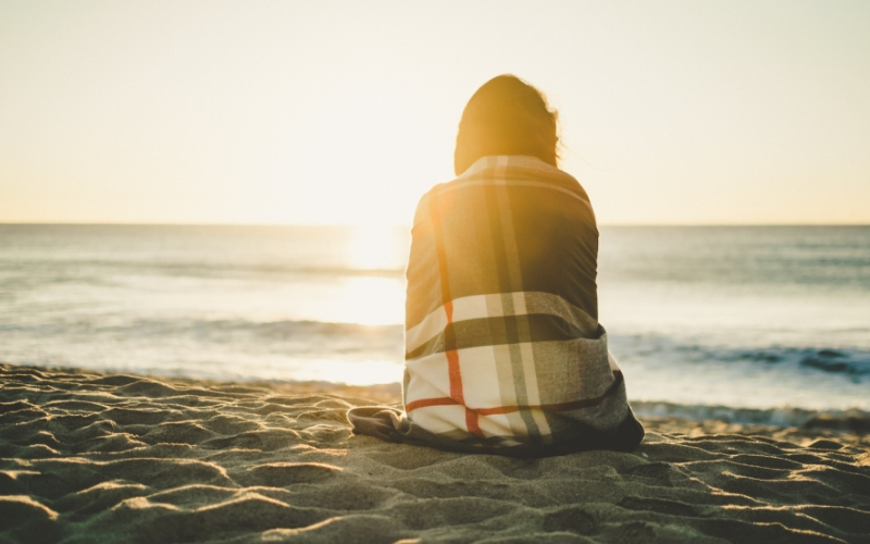 woman sitting on the beach at sunrise wrapped in a plaid blanket