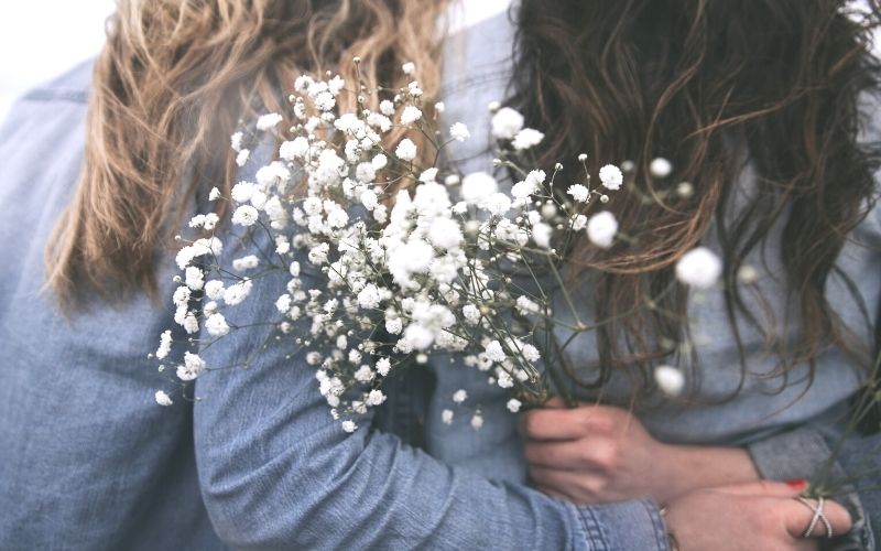 picture of two women in denim shirts, from the back, arms around each other, with a bouquet of white flowers