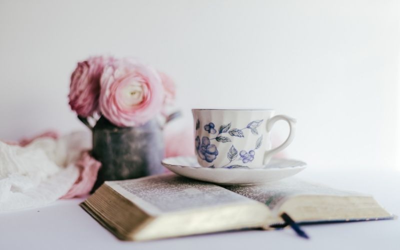 Open Bible with china teacup and saucer sitting on top, pink roses in background.