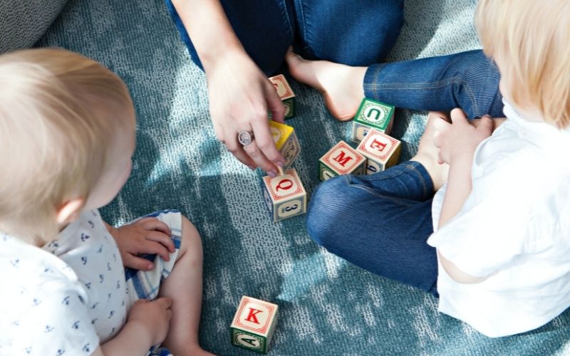 Two small blond children playing with blocks, with their mother or caregiver