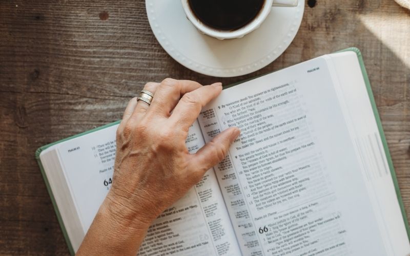 Woman's hand resting on a Bible on top of a wooden table, with a cup of coffee next to it.