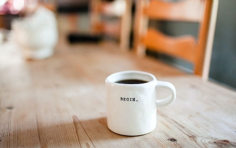 Coffee on a wooden dining room table, in a white mug with the word 'BEGIN.'