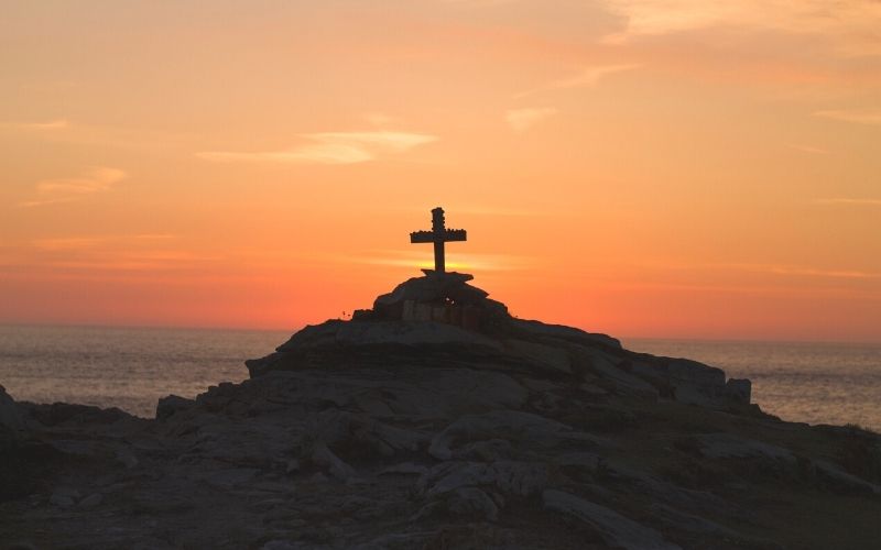 wooden cross on a rocky hill by the ocean at sunset